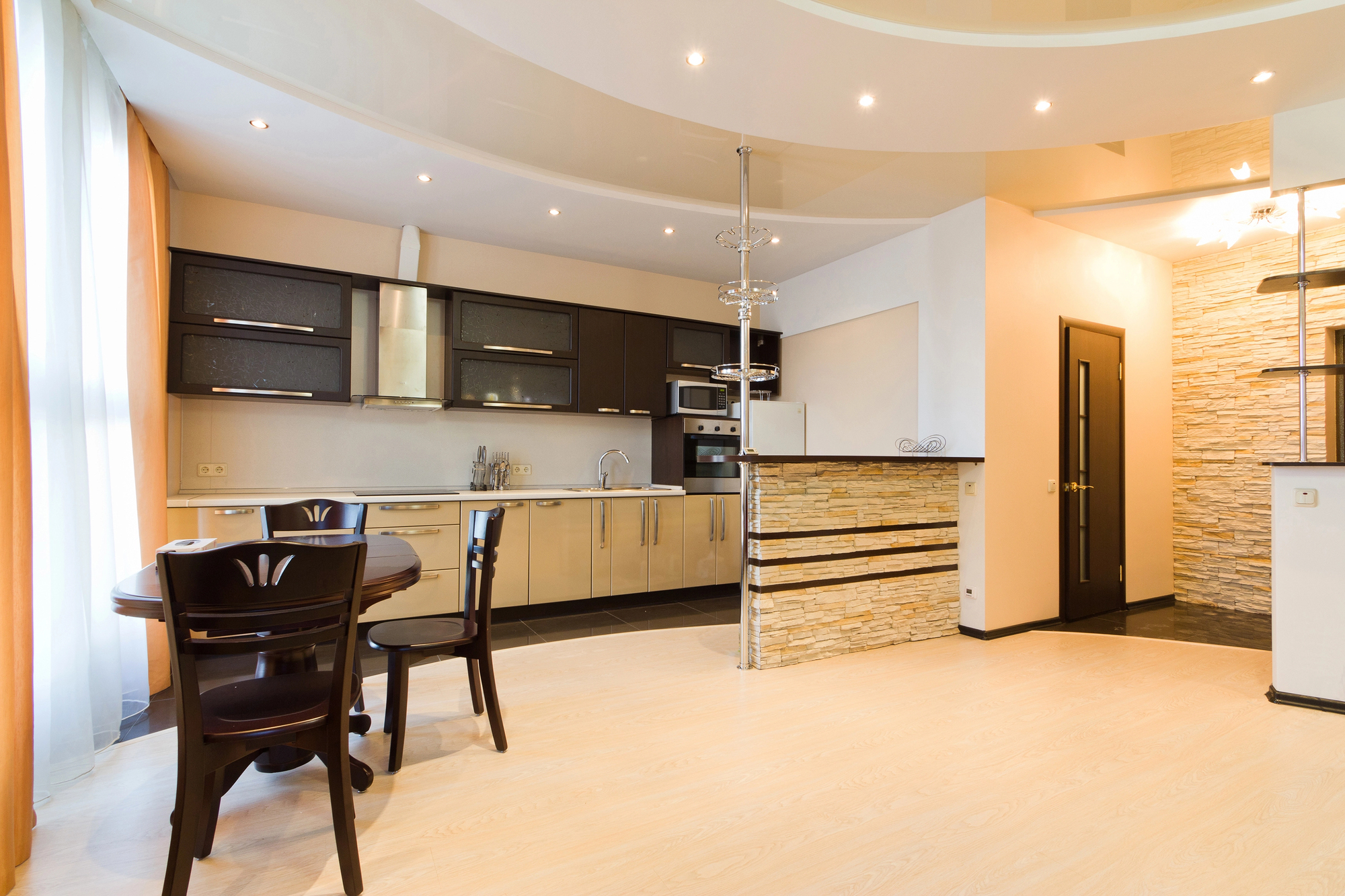 Modern open-plan kitchen and dining area with light wood floors, black and beige cabinetry, a round dining table, stone-accented bar, and recessed ceiling lights—showcasing expert property refurbishments by a skilled builder.