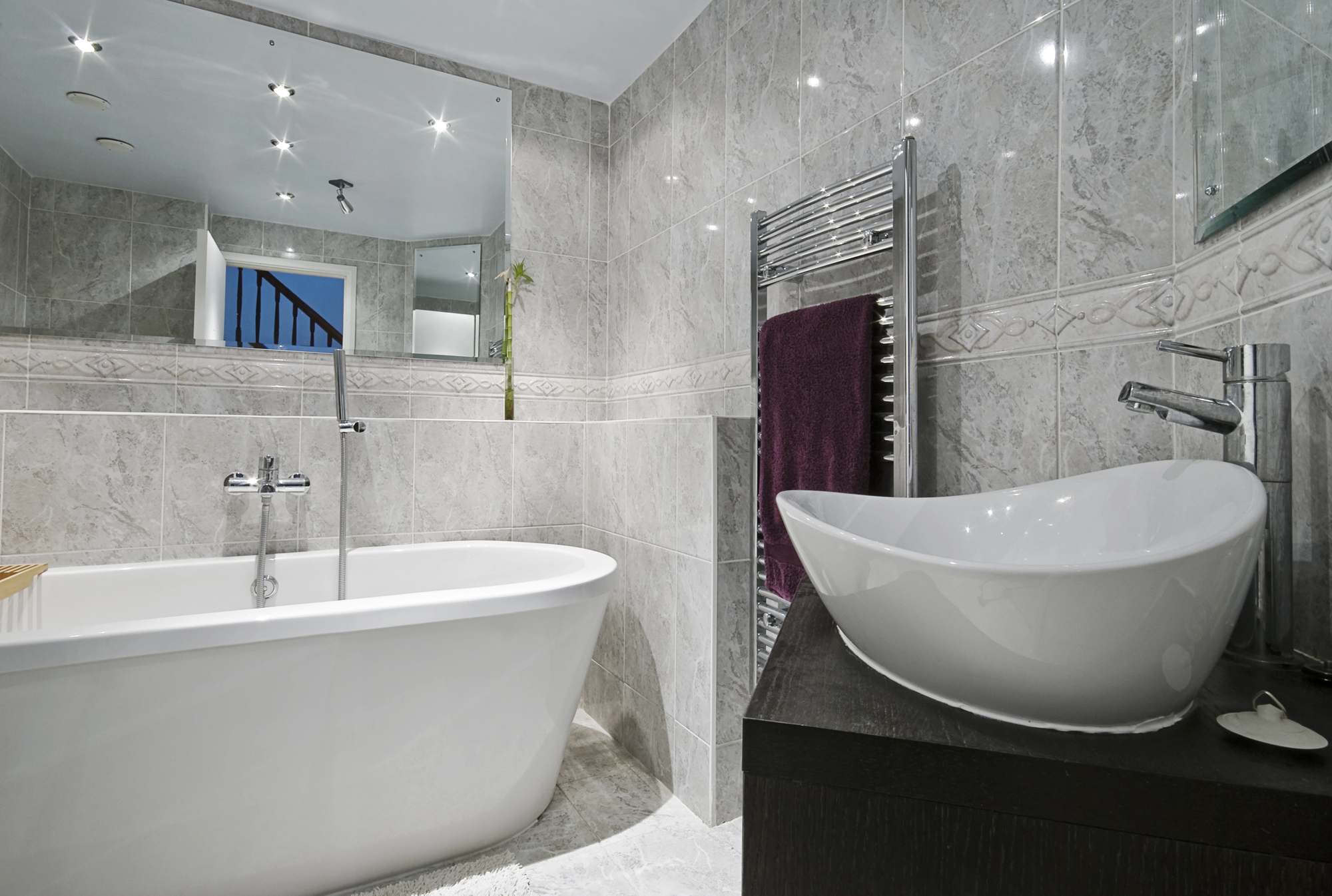 Modern bathroom with a white freestanding bathtub, vessel sink on a dark vanity, heated towel rail with a purple towel, large wall mirror, and gray marble tiles—precision that reflects expert builder and building maintenance standards.