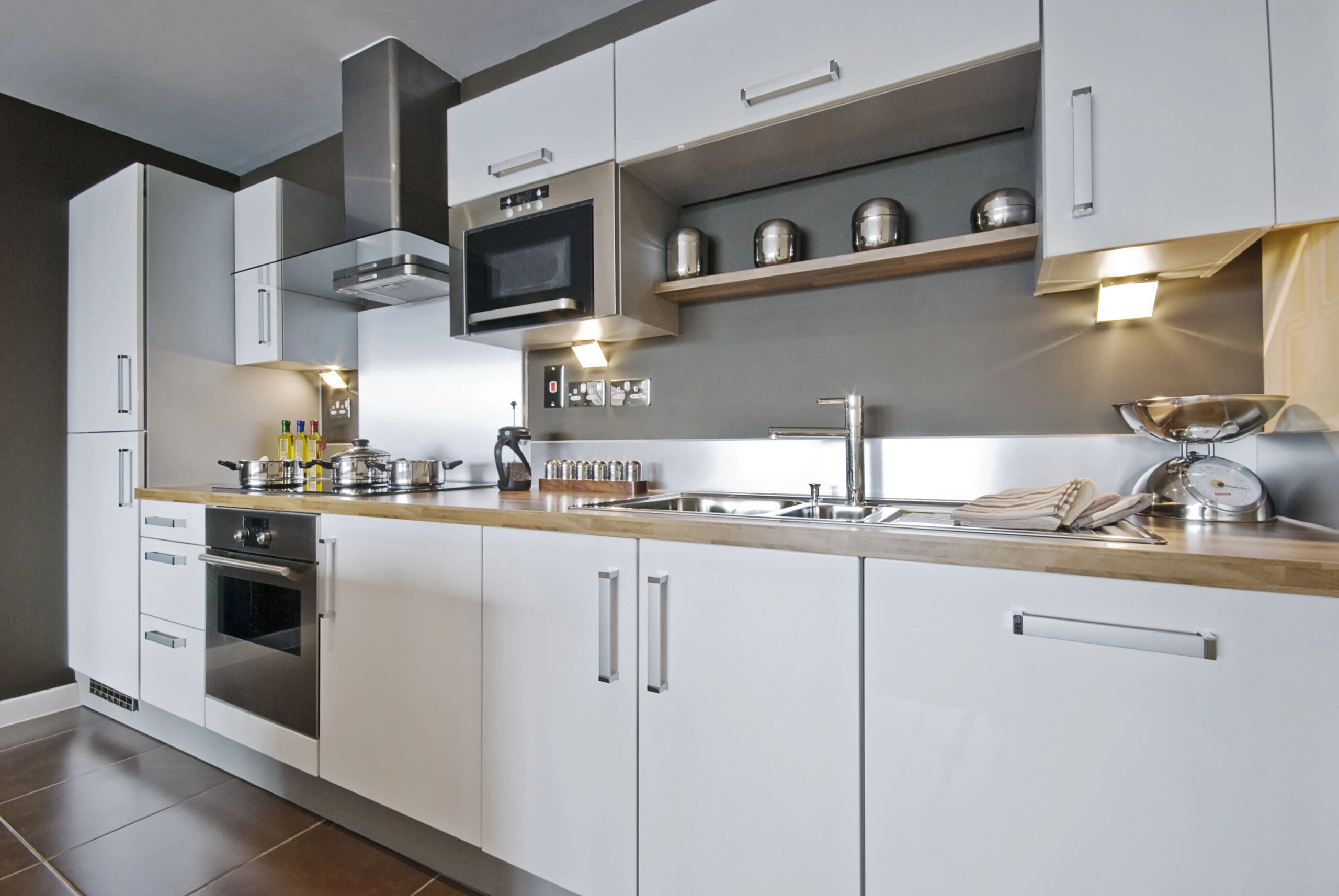 A modern kitchen with white cabinets, stainless steel appliances, a built-in oven, microwave, sink, and a wooden countertop. Shelves hold metal canisters and spices; under-cabinet lights brighten this sleek Kitchen Installation.