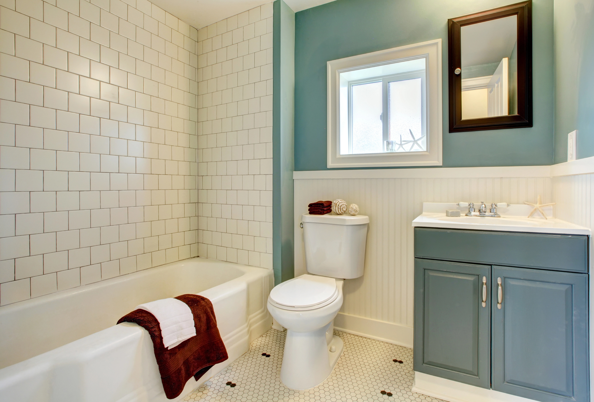 Bathroom with white tile bathtub and shower, white toilet, blue vanity with sink, mirror, and a window—ideal after recent property refurbishments. Towels are folded on the tub and vanity. Walls are blue and white with hexagonal floor tiles.
