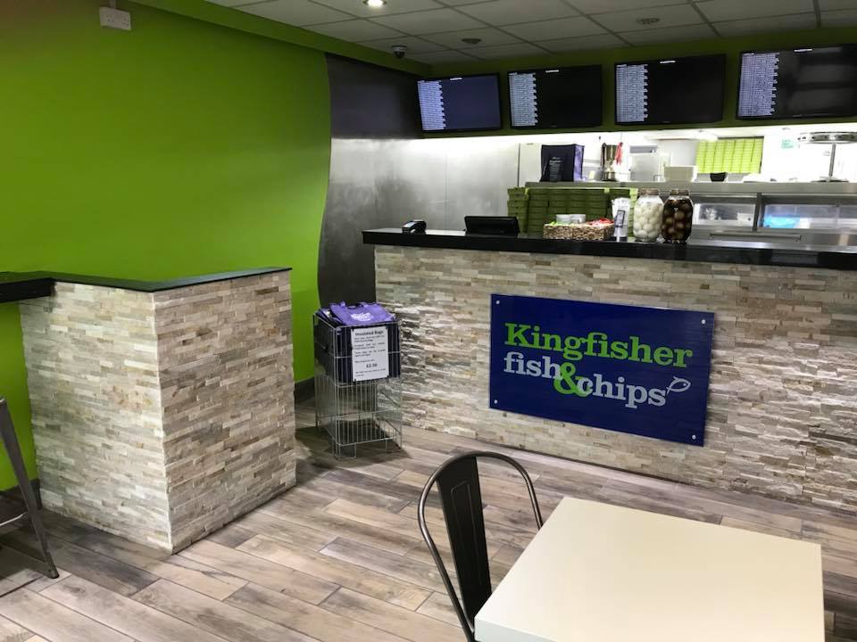 Interior of a modern fish and chips shop with light wood floors, green walls, and a counter with a "Kingfisher fish & chips" sign—showcasing recent building maintenance updates, including new bathrooms. Digital menu screens hang above; table and chairs in the foreground.